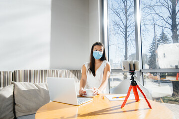 A young girl is sitting in a cafe in a mask and leads a video blog. Communication to the camera.