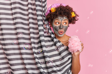 Surprised dark skinned young woman with curly hair applies beauty clay mask on face looks aside holds shower sponge enjoys showering poses in douche hides behind curtain soap bubbles around.