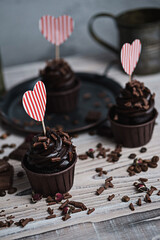 Several muffins or cupcakes with chocolate shaped cream at white table. A card in the form of a heart for St. Valentine's Day. A woman's hand crumbles grated chocolate onto a cake.