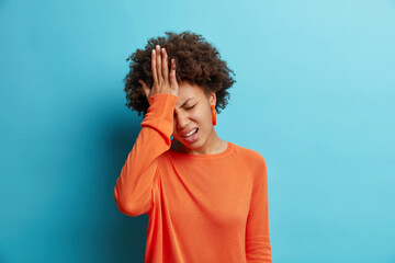 Fototapeta na wymiar Frustrated dark skinned young woman keeps hand on forehead regrets wrong doing feels stressed dressed in orange sweater isolated over blue background. Upset Afro American girl facepalming indoor
