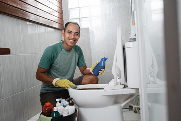 smiling male looks to camera hold sponge and a cleaning fluid bottle when cleaning the dirty toilet in the bathroom