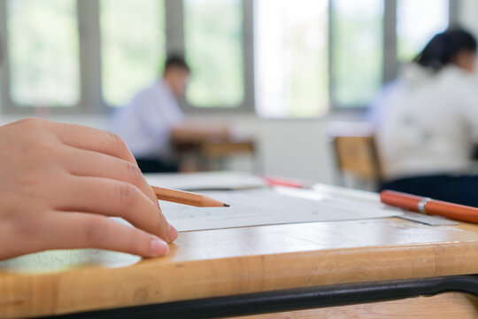 High school or university student reading question exam test and using pencil writing on paper answer sheet, lecture chair doing final exam attending in examination classroomwith student in uniform