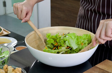 Fototapeta na wymiar The housewife dressed in an apron, mix the vegetables in a salad bowl together with a wooden ladle. Morning atmosphere in a modern kitchen.