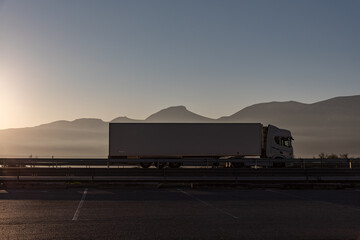 Fototapeta na wymiar Truck with refrigerated semi-trailer driving on a highway with mountains in the background, side view and the sun shining from behind.