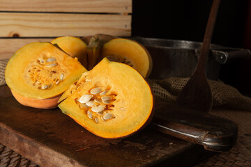Pumpkin, rustic arrangement with pumpkin cut into pieces, knife, board, wooden spoon, iron pot on a table, with rustic fabric and wood, black background, selective focus.