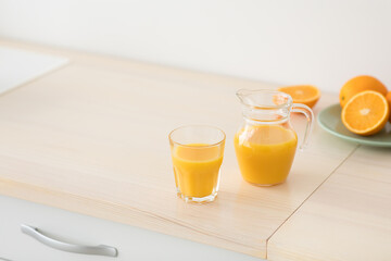 Freshly squeezed orange juice in glass and jug, fruits on plate on white furniture in modern minimalist kitchen