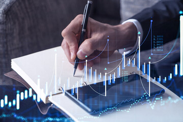 A trader in formal wear writing down some quotes to research stock market trends using smart phone for right investment solutions. Wealth management concept. Hologram Forex chart over close up shot.
