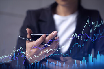 A woman trader holding in the hands a smart phone and researching stock market to proceed right investment solutions. Internet trading and wealth management concept. Hologram Forex chart.