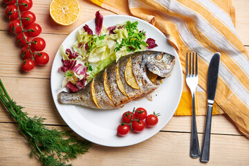 Tasty grilled dorado fish with lemons and herbs served on the plate with tomatoes and salad. Top view.