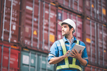 Engineer or foreman holding tablet and wears PPE looking at left side to checking inventory or job details with cargo container background. Engineering site and working with technology concept.