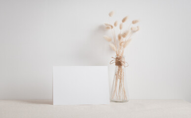 Blank white greeting card mock up.decoration with dried Lagurus ovatus flowers composition in modern glass vase  on  beige table and cement wall background