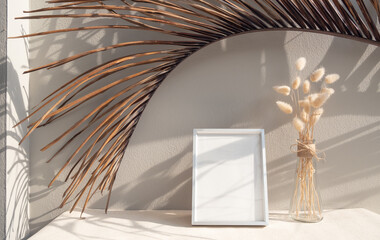 Blank mockup picture frame,coconut palm leaf and dried flower in glass vase on beige table and cement wall background