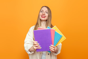 Caucasian blonde student girl in beige jacket holds four books in multi-colored covers smiling isolated over orange background in studio. English language school, education concept