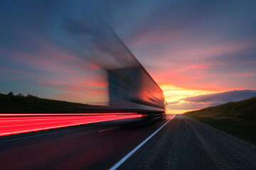 Motion blur. A large truck is driving along the highway at high speed. Sky with bright red clouds. Delivery of cargo. - obrazy, fototapety, plakaty