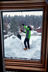 Snow removal on the roof of a country house. Boy in green jacket with shovel throws snow off the...
