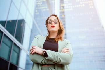 Caucasian woman, with round nerdy glasses. With arms folded, with a serious expression, in a background of glass buildings. Job, economy, companies, boss woman and businesswoman concept. Copyspace