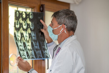 Doctor wearing face mask looking at x-ray backlit at the room window