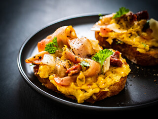 Delicious breakfast - scrambled eggs with bacon and champignons and sun-dried tomatoes on toasted bread on black table
