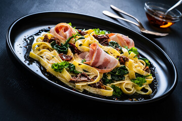 Tagliatelle with bacon, sun-dried tomatoes and spinach in alfredo sauce on black wooden table 
