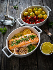 Roasted salmon steak with roast potatoes and tomatoes served in ceramic pan on wooden table
