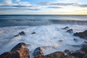 A closeup shot of rocks in the wavy sea under a sunset sky