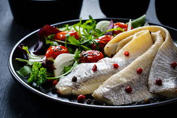 Marinated herrings with roast cherry tomatoes and fresh vegetable leaves on black table
