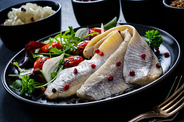 Marinated herrings with roast cherry tomatoes and fresh vegetable leaves on black table
