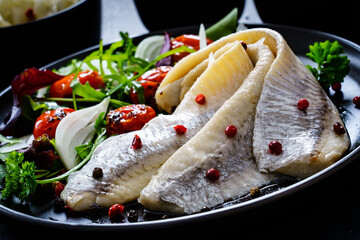 Marinated herrings with roast cherry tomatoes and fresh vegetable leaves on black table
