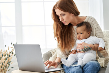 Busy young single parent caucasian mother using laptop computer distance working online from home office having video call virtual meeting with little cute african american infant baby girl daughter.