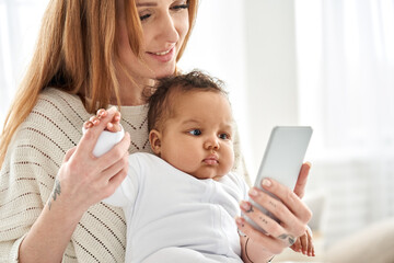 Happy young caucasian mother holding cute african american baby girl looking at smartphone at home....