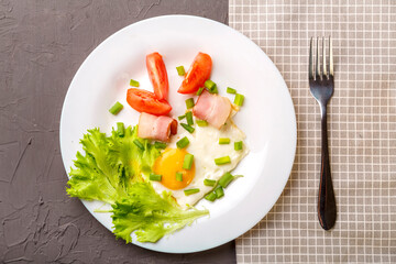 Scrambled eggs with bacon garnished with tomato and green onion on a gray concrete background on a napkin next to a fork.