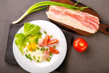 Scrambled eggs with bacon, garnished with tomato and green onion and on a gray concrete background on a wooden stand next to chopped bacon and tomato.