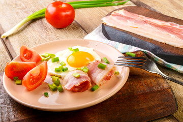 Fried eggs with bacon with tomato and green onion and on a wooden table in a plate next to chopped bacon and tomatoes.