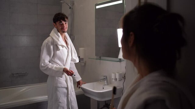 A young European in the bathroom is in a robe and brushes his teeth, his girlfriend watches as the guy takes care of the tooth enamel.