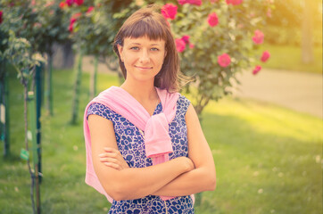Naklejka premium Young beautiful caucasian girl with dark hair and flowered shirt looking at camera, posing, crossing arms and smile in garden with flowers and green trees in sunny summer day with sun flare backlit