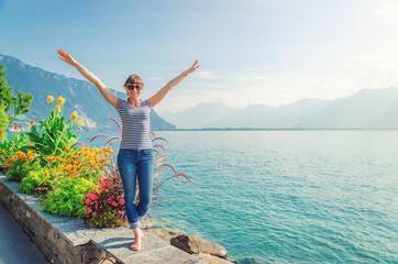 Fototapeta na wymiar Young beautiful girl traveler with striped shirt and jeans posing, raise hands and smile on embankment of Lake Leman Geneva in Montreux town of Swiss Riviera, mountains Alps background, Switzerland