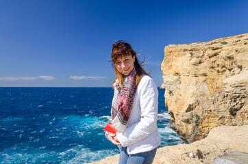 Young woman traveler with white jacket and headscarf looking at camera and smile on stone rocks of Dwejra Bay with collapsed Azure Window in Gozo island of Mediterranean sea, Malta in summer day