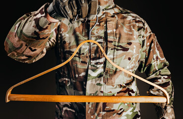 Photo of soldier in camouflaged uniform and tactical gloves holding wooden shirt hanger on black background.