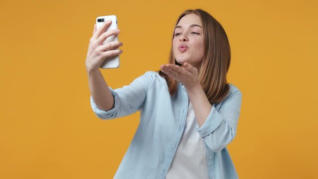 Cheerful young woman 20s in blue shirt isolated on yellow background studio. People lifestyle concept. Doing selfie shot on mobile phone showing victory sign blowing sending air kiss put hand on cheek