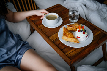 Female enjoying a cup of coffee with delicious pastries on a board on the bed