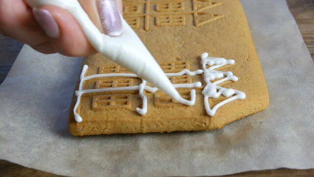 Brown gingerbread frosting. Female chef's hands decorate with white sugar icing and pastry bag a gingerbread Christmas house in the home kitchen. New year traditions.