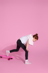 Stylish little child girl is learning to skateboard for the first time over pink background.