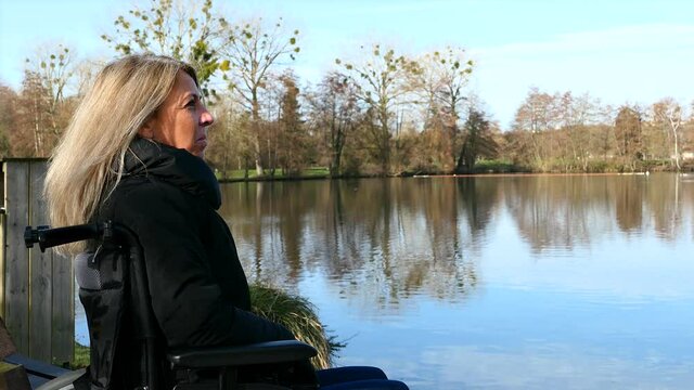 Concept of disabled person. A woman in a wheelchair outside in the nature in front of a lake. 