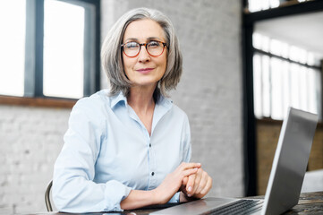Intelligent mature senior female business owner in glasses looking at camera, sitting at the desk...