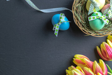 Easter background blue. Colorful egg with tape ribbon, spring tulips on dark rough stone background in Happy Easter decoration. Flat lay, top view.