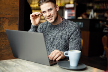 Confident young man in smart casual clothes and glasses, working on laptop. Beautiful smiling male in cafe drink coffee. Blogger influencer looking at camera make video conference call recording.