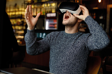 Happy man in glasses of virtual reality in cafe. Smiling male using VR helmet. Augmented reality game, future technology, AI concept. 