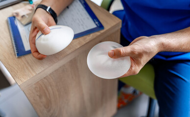 Plastic surgeon man is holding different size silicon breast implants in clinic room interior....