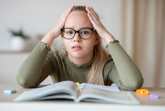 Upset teen girl in glasses sitting at table with book