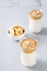 Quarantine trendy cuisine. Two cups with dalgona coffee and banana chips in a bowl on a gray background. Vertical view
